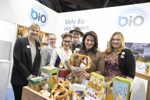 Gruppe von Personen an einem Messestand mit Bio-Produkten aus Bayern, darunter Brezeln, Getränke und Lebensmittel.